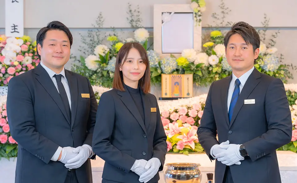 世田谷区家族葬 はばたきグループの男性スタッフ2名と女性スタッフ1名が葬儀に使用する生花祭壇の前で、そっと手を組んでご遺族をお待ちしている様子